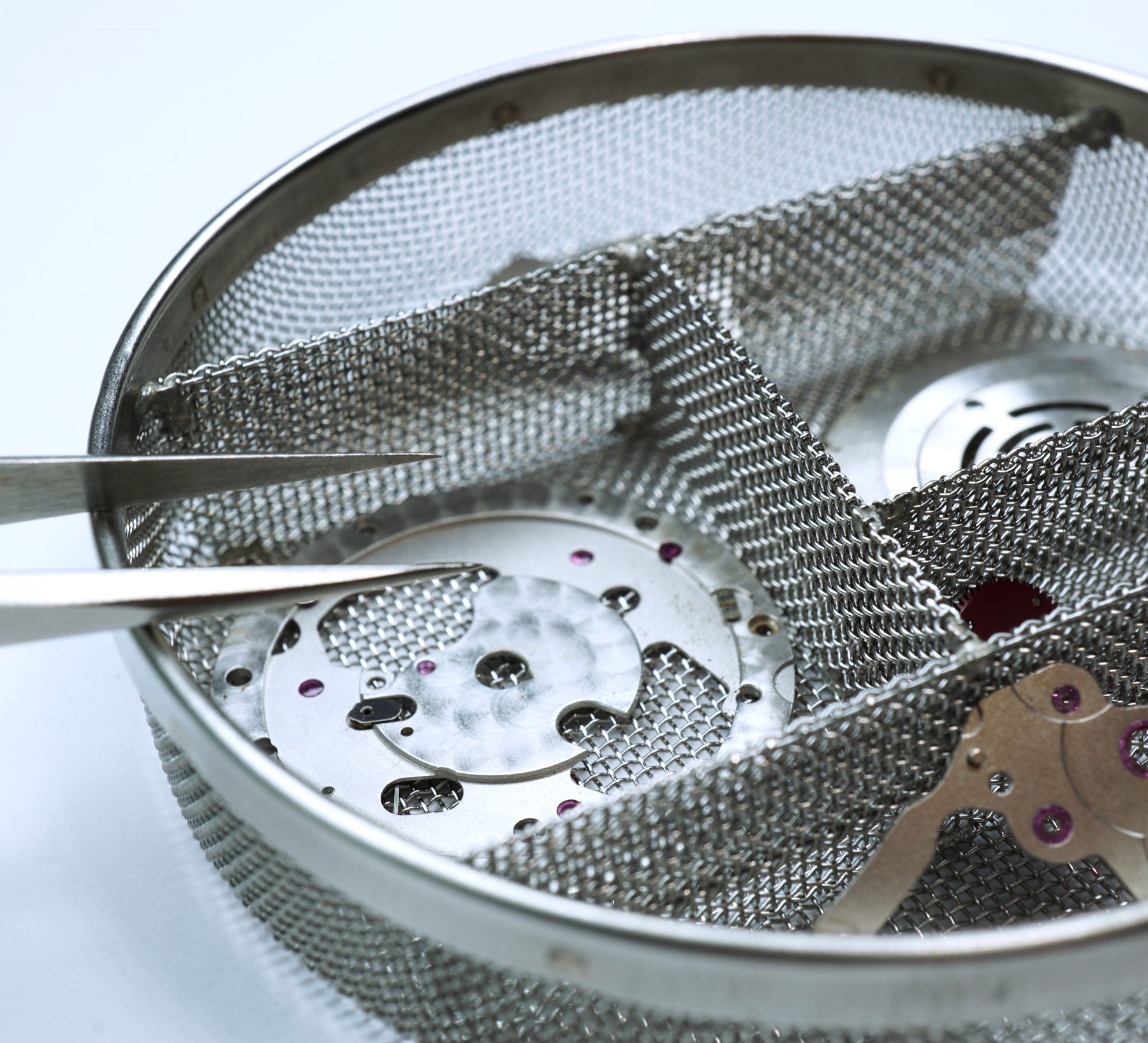 Watch movement components in a mesh cleaning basket, held with tweezers